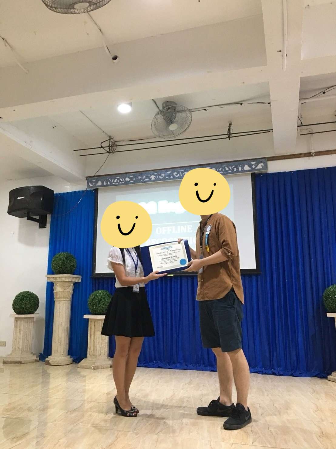 QQEnglishのロゴの前で修了証を受け取ったときの写真